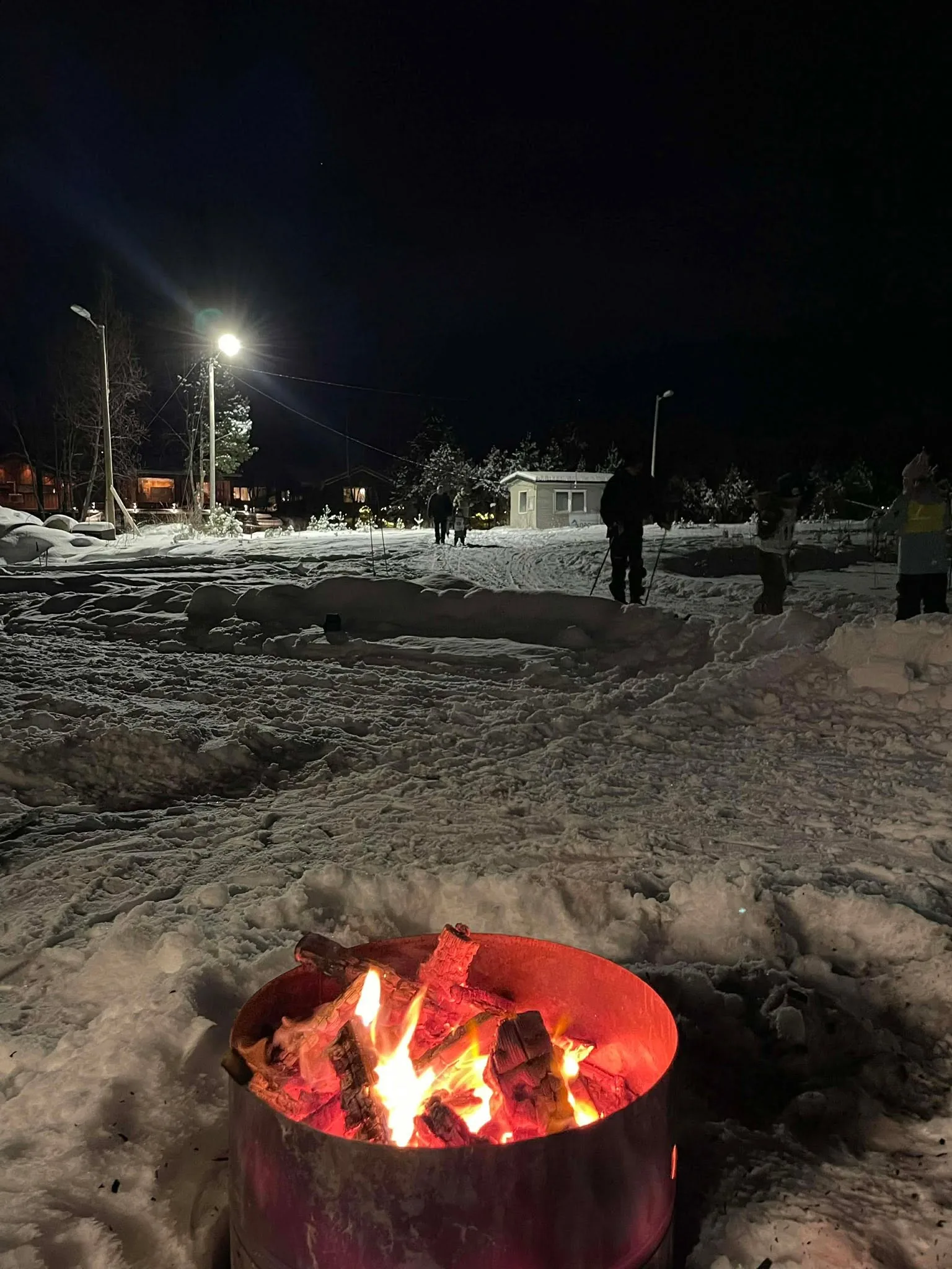 Bålpanne som brenner i snøen utenfor Idrettsheimen om kvelden